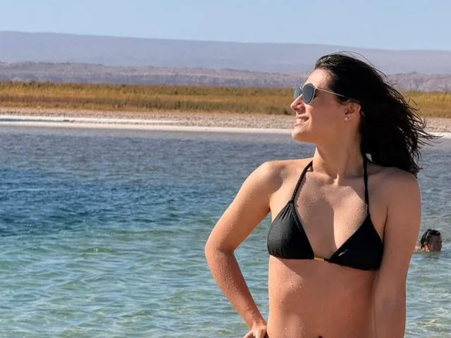 A woman in a black bikini poses by a clear, turquoise lagoon.