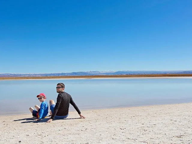 Un homme et un enfant sont assis sur la rive sablonneuse d'une grande lagune peu profonde.