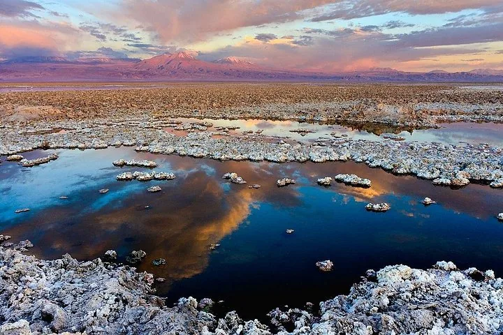 Atardecer sobre un salar con el agua reflejando el cielo colorido y las montañas.