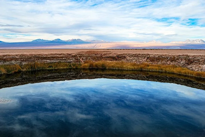 Une doline parfaitement immobile reflète le ciel nuageux et une chaîne de montagnes lointaine.